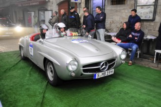 Silver 190 SL Roadster with helmeted occupants slowly across the green carpet, watched by