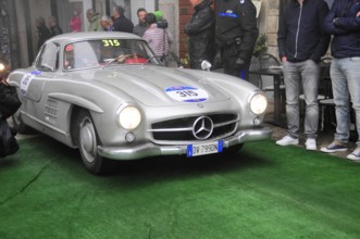 Silberner 300 SL drives through the old town with light over green carpet, surrounded by spectators