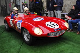 Red racing barchetta with start number 16 glides across the green carpet, two drivers wearing