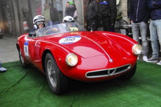 Red roadster with two helmeted drivers rolls across the green carpet at the rally, flanked by