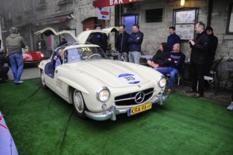 Creamy white 300 SL with open double doors poses on the green carpet, surrounded by smiling