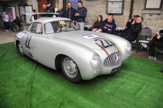 Silver Mercedes gullwing door with race number in a narrow alley at a classic car rally, spectators