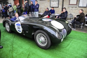 Black jaguar roadster rolls across green carpet during a historic rally, spectators sitting close