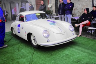 White Porsche Coupé at a classic car rally on green carpet, spectators lean against the stone wall,
