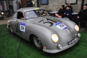 Silver Porsche Coupé rolls across green carpet in front of spectators on a stone wall at a historic