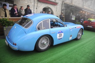Light blue vintage racing car with start number 201 rolls across green carpet through a historic