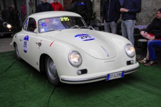 White Porsche 356 with glowing headlights drives across green carpet during a vintage car rally,
