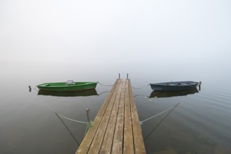 Two boats are moored on a wooden dock in a foggy lake, quiet and peaceful atmosphere, Hopfensee,
