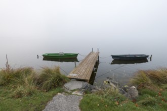 Two boats and a jetty on foggy lakeside with grass and stones, Hopfensee, Ostallgäu, Bavaria,