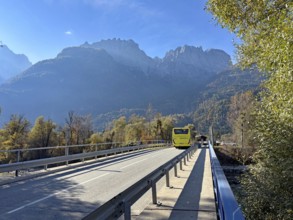 A yellow bus crosses a bridge under a blue sky, surrounded by autumnal mountains, Lienz Dolomitem,