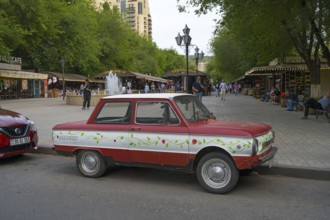 Red retro car parked in front of busy market square with green trees, SAS-968, Zaporoshez, Yerevan,