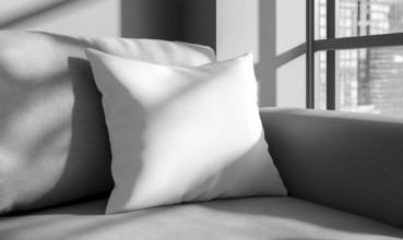 A white pillow sits on a gray couch in front of a window. The pillow is placed on the couch in such