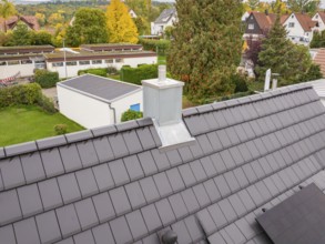 Roof with anthracite-colored tiles and modern chimney, behind it quiet residential area with autumn