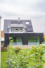 Front view of a house with solar panels and black extension with glass front, well-kept garden and
