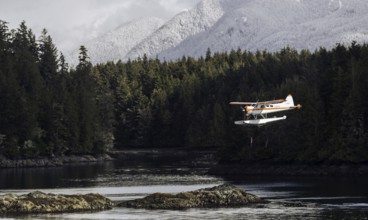 A seaplane flies above rocky shores and dense forest with snow-capped mountains in the background