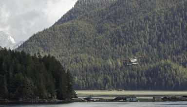 A seaplane flies over a tranquil lake surrounded by dense forest and mountains under an overcast