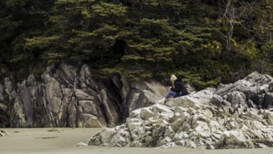 A person sits on rocky coastline surrounded by trees under a cloudy sky in Tofino, British