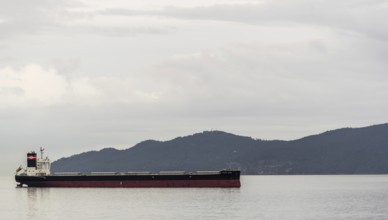 A solitary cargo ship sails by a mountainous backdrop under cloudy skies in Vancouver, British