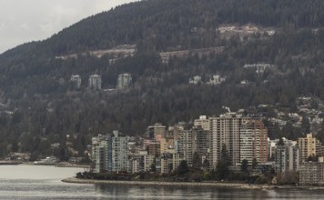 Urban high-rise buildings set against a backdrop of forest and mountains on an overcast day in