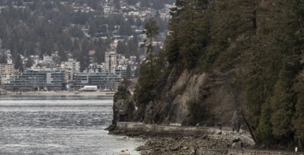 Rocks and trees line a coastal path in Stanley Park with urban buildings in the distance in
