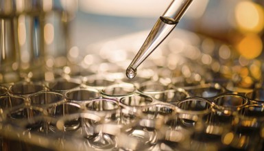 A warm-lit droplet hangs from a glass pipette above gleaming wells, evoking meticulous care in a