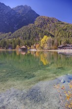 Emerald green, clear shore water with visible stones, colorful autumn trees and a boathouse are