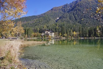 Wide shore with shallow, crystal-clear water, opposite is a villa in the sunshine in autumn forest,