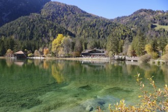 Clear mountain lake with emerald green water, autumnal forest and hut on the shore under blue sky,