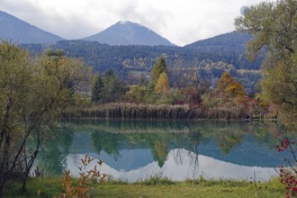 Quiet lake with turquoise-green water, autumnal shore and mountain scenery under cloudy sky, Drau,