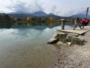 A bicycle is leaning on a footbridge over a still lake, mountains and clouds are reflected in the