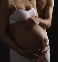 Pregnant young long haired woman staying close to her husband both tenderly looking at her belly