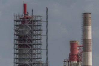 Workers work on a large chimney covered with scaffolding. Two chimneys are visible and the sky is