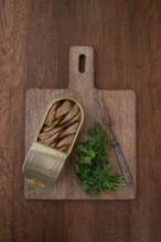 An open can of sprats, appetizer, on a wooden cutting board, with a fork and herbs, top view