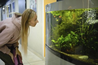 A woman leans forward, observing vibrant fish swimming in a large aquarium. The setting is bright