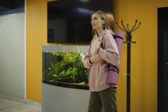 A woman stands in a fitness studio holding a yoga mat. She wears a cozy hoodie and looks ready for
