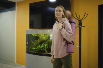 A woman in a pink hoodie stands in a fitness studio, holding a yoga mat. An aquarium with aquatic