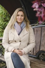A woman sits calmly on a wooden bench, wearing a beige trench coat and gray scarf. She exudes a