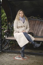 A woman with long hair sits on a bench in a city park. She wears a beige coat, gray scarf, and