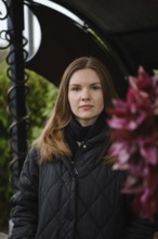 A young woman with long hair poses confidently near vibrant flowers in a garden. The scene captures
