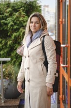 A woman stands outside a contemporary building, dressed in a light coat and carrying a backpack.