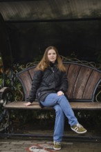 A young woman sits calmly on a wooden bench in a park surrounded by autumn foliage. She wears a