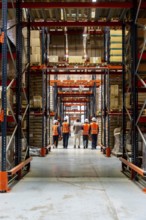 Workers in hard hats and reflective vests walk down a high rack warehouse aisle filled with stacked