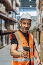 Male warehouse worker in orange safety vest and white hard hat scans inventory with a handheld