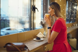 A woman in a red dress sits by the window of a cafe, sipping a warm drink and enjoying a dessert.