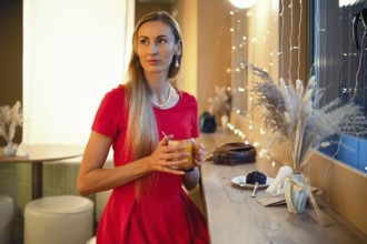 A woman with long hair sits at a bar in a cozy cafe, holding an orange drink. She looks thoughtful