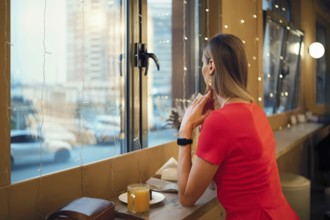 Rear view of a woman dressed in a vibrant red dress sitting near a window in a cozy cafe, looking