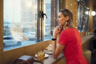 A woman sits quietly in a cafe, gazing out the window at the city skyline. Soft lights surround her