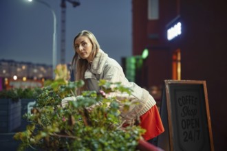 A woman leaning on the railing decorated with the plants outside a coffee shop in the evening. The