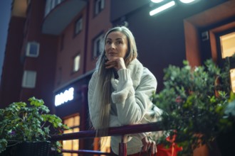 A woman stands thoughtfully on a balcony railing, dressed warmly for the evening chill. The city