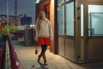 A woman strolls outside a coffee shop in the evening. She is dressed in a bright red dress, brown